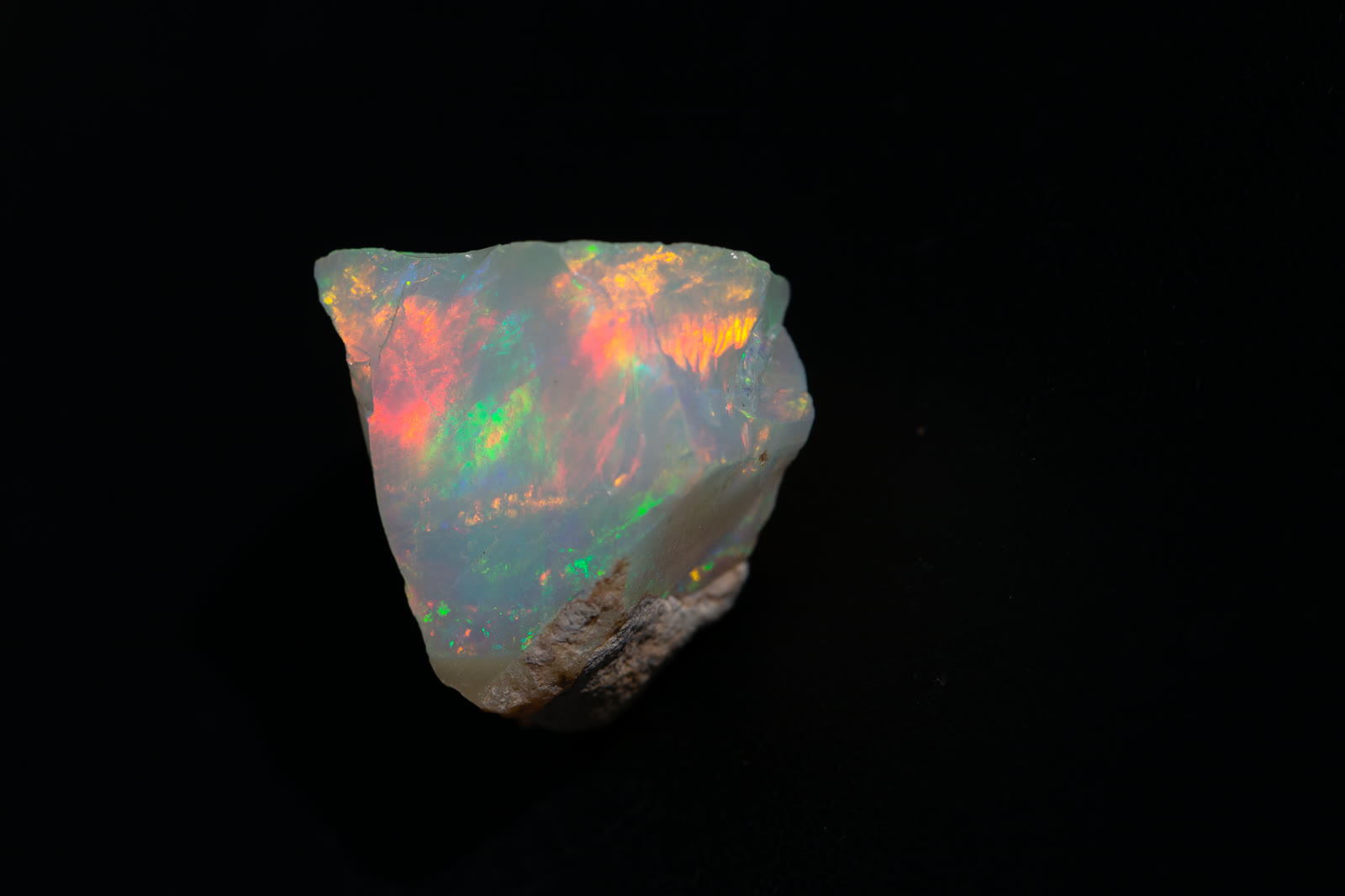 A 25.8-carat piece of rough Ethiopian Welo opal on black — rolling red, orange, and green flash across a crystal body with matrix at the base.