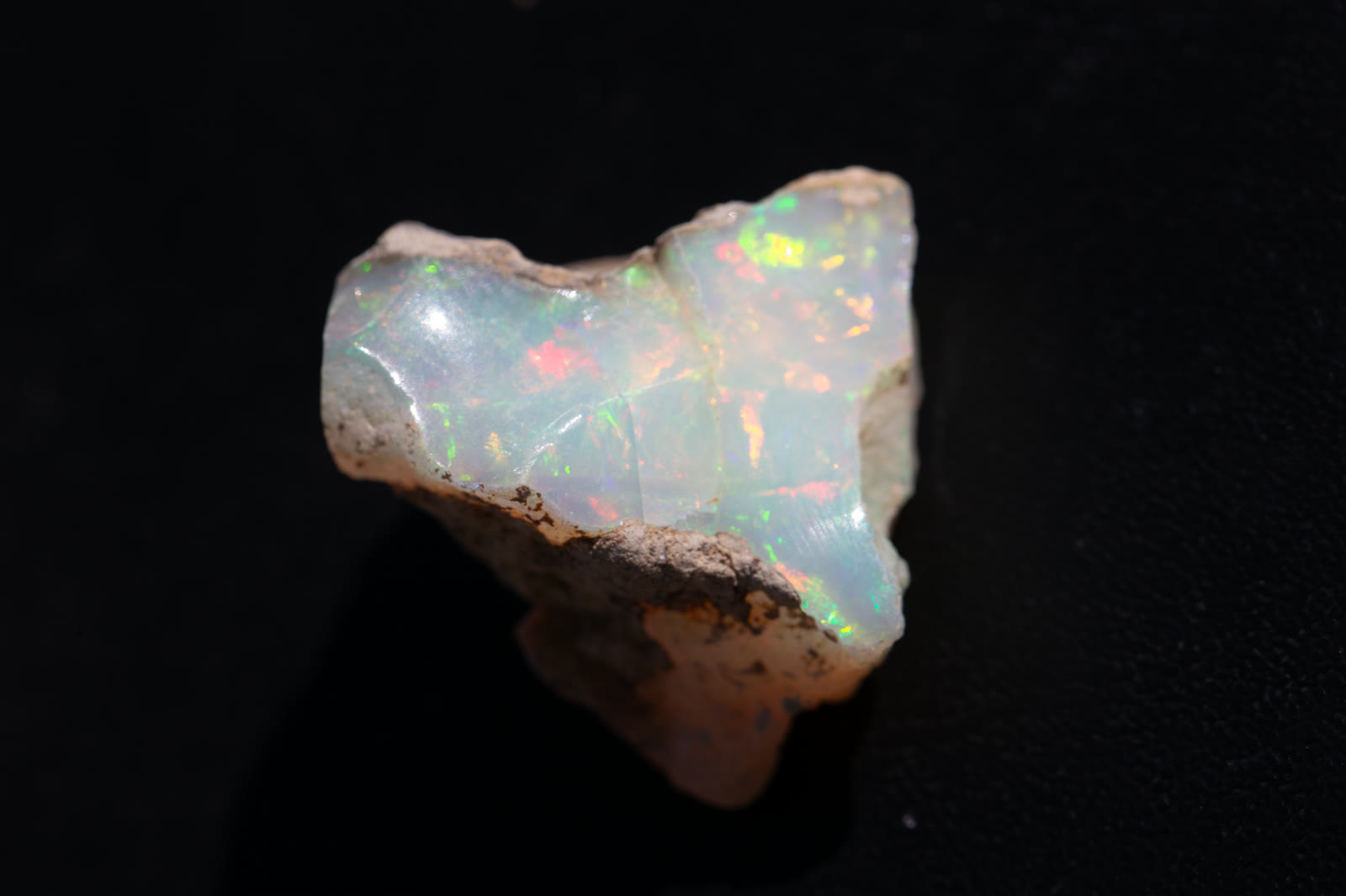 A 25.95-carat piece of rough Ethiopian Welo on black — lighter crystal body, soft pinfire of red and green across the face.