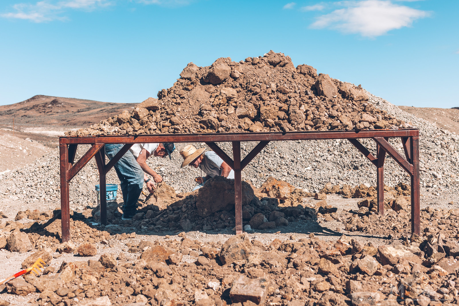 Two diggers working through a pile of dirt and rock heaped on a steel table at Rainbow Ridge, high desert hills behind.