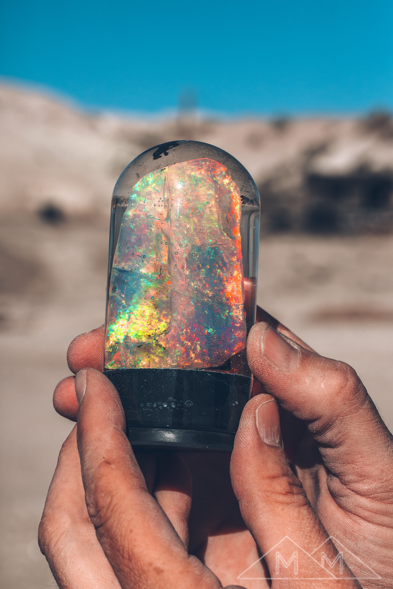 Two dusty hands holding a water-filled display dome up against the desert sky, a large crystal opal glowing inside.