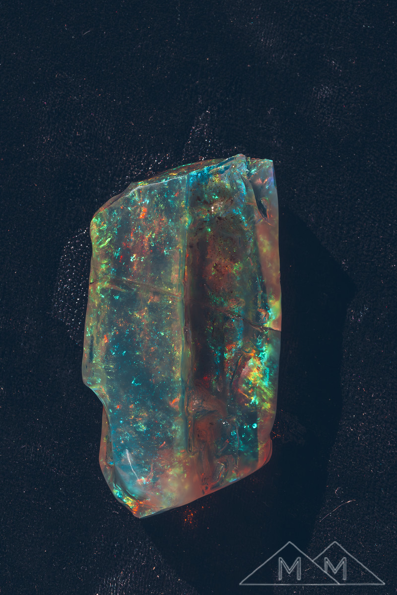 The crystal opal photographed on black velvet, teal and red fire across the full face.