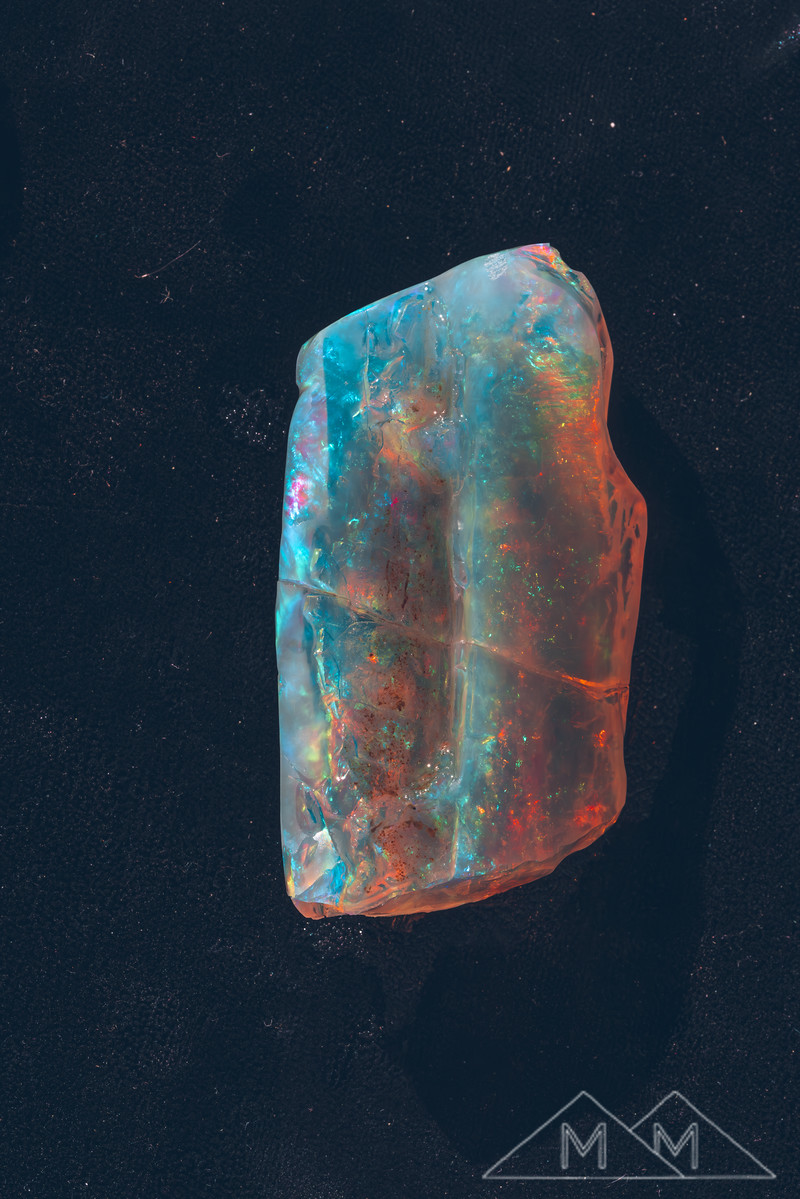 The crystal opal on black velvet from another angle, blue at the top falling into deep orange fire.