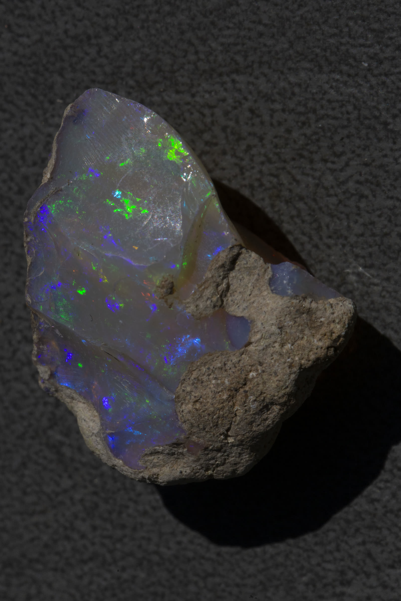 Rough Ethiopian Welo crystal opal with vivid blue and green play-of-color, glassy transparent body, sandy matrix still attached.