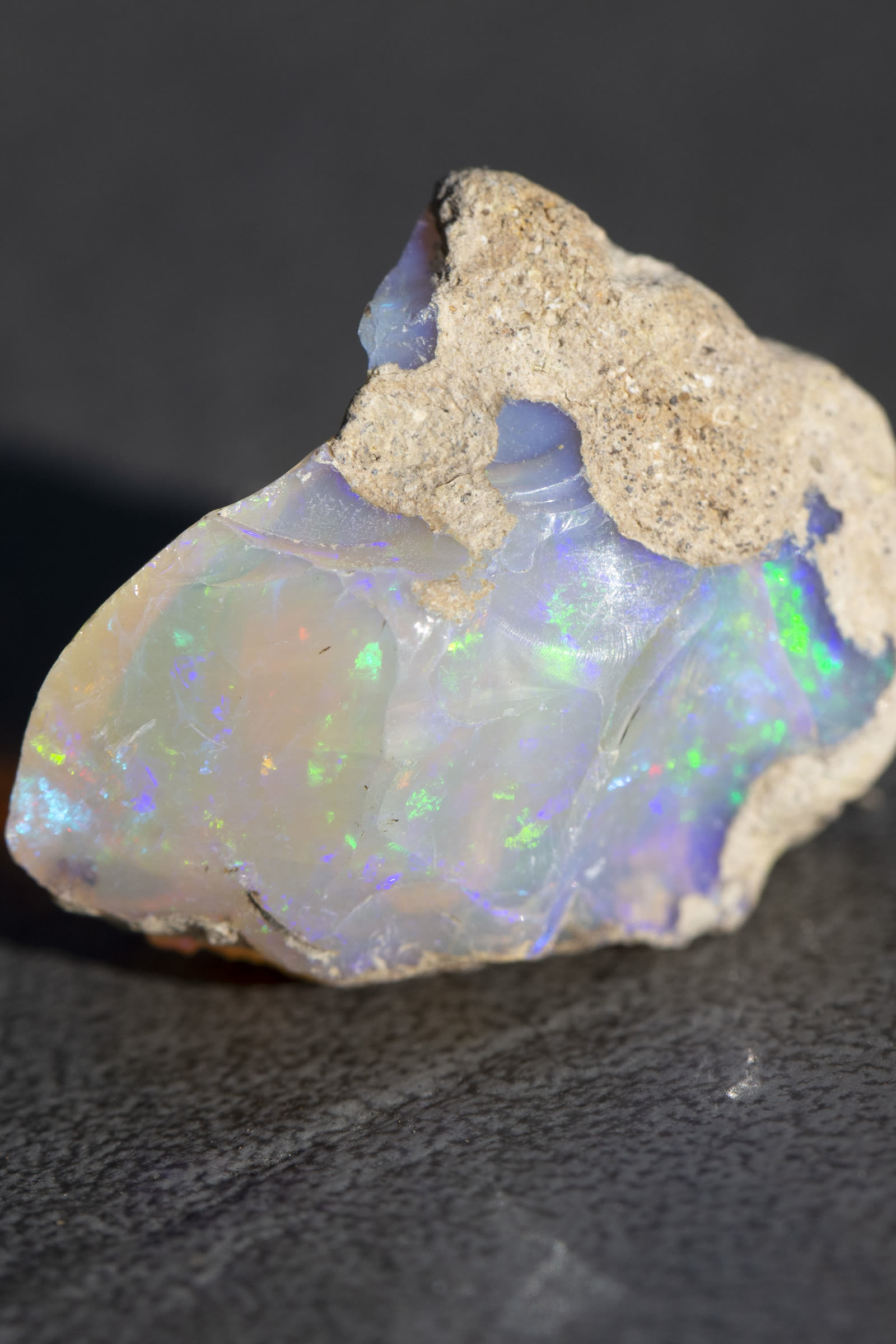 Ethiopian Welo crystal opal rough with a translucent milky-lavender body, green and blue play-of-color showing through the stone, sandy matrix still attached at top.