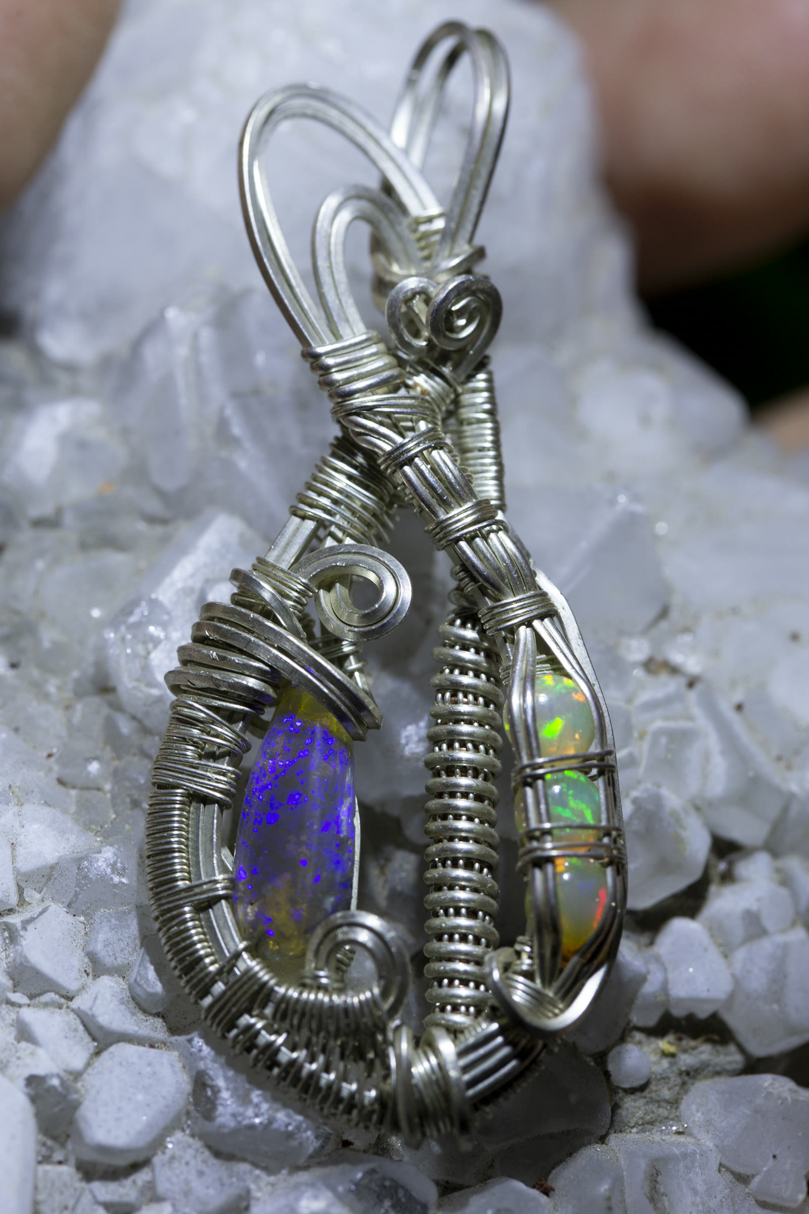 A wire-wrap pendant holding two opals in a single silver wire frame — a dark blue opal on the left and a lighter crystal opal with pinfire on the right, set in coiled fine silver wire against a quartz crystal.